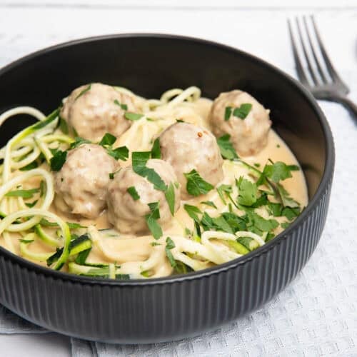 Square image of swedish meatballs with vegetable zoodles in a black bowl on a white cloth