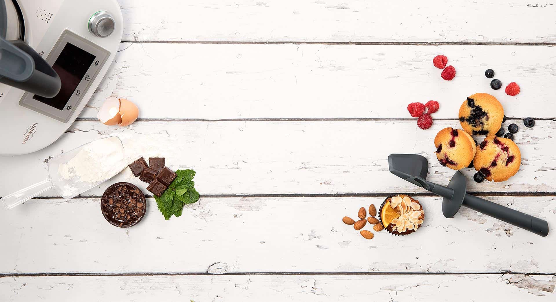 Thermomix on a wooden background with a spatula, ingredients and cakes