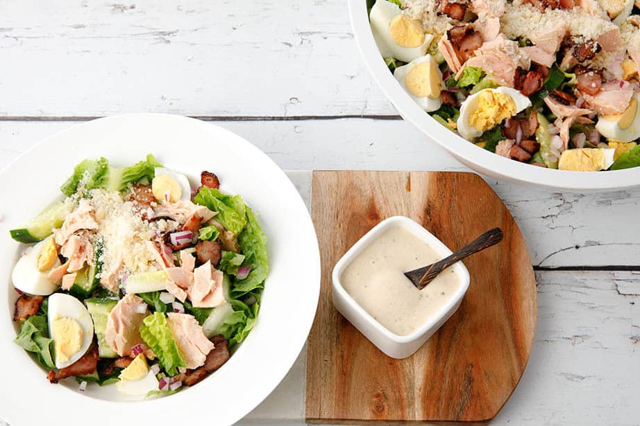 Overhead shot, white bowl of Caesar Salad with dressing on the side served from a large salad bowl