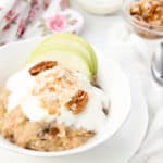 White bowl of Apple pie porridge on a white and floral background with apple, yoghurt and nut toppings.