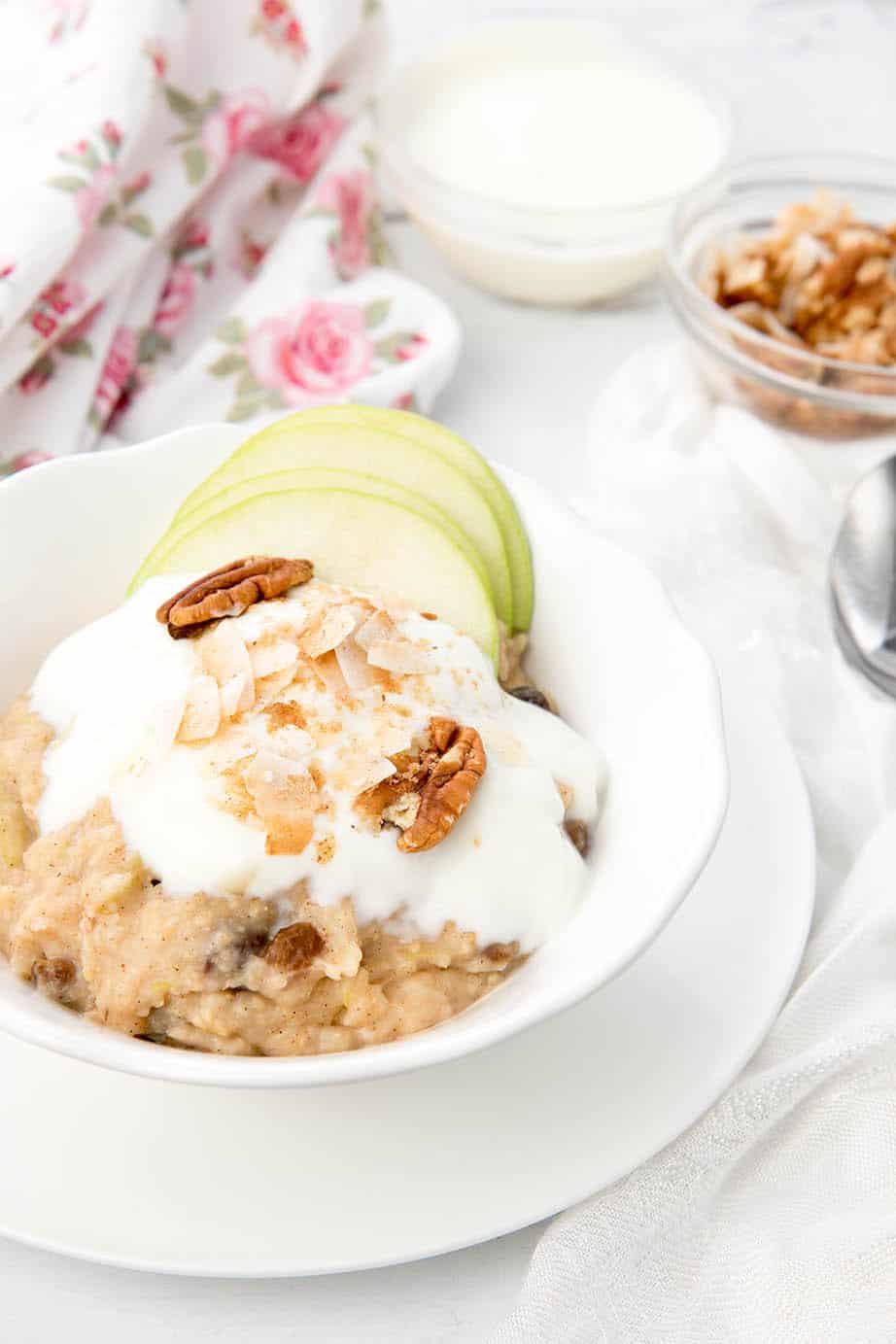 White bowl of Apple pie porridge on a white and floral background with apple, yoghurt and nut toppings.