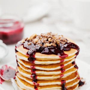 Pancake recipe showing a light coloured table setting with a stack of pancakes with dripping red berry sauce