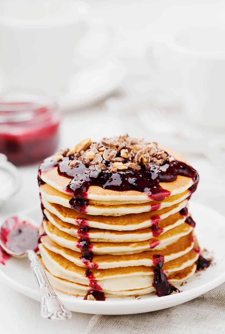 Pancakes on a light coloured table setting with a stack of pancakes with dripping red berry sauce 