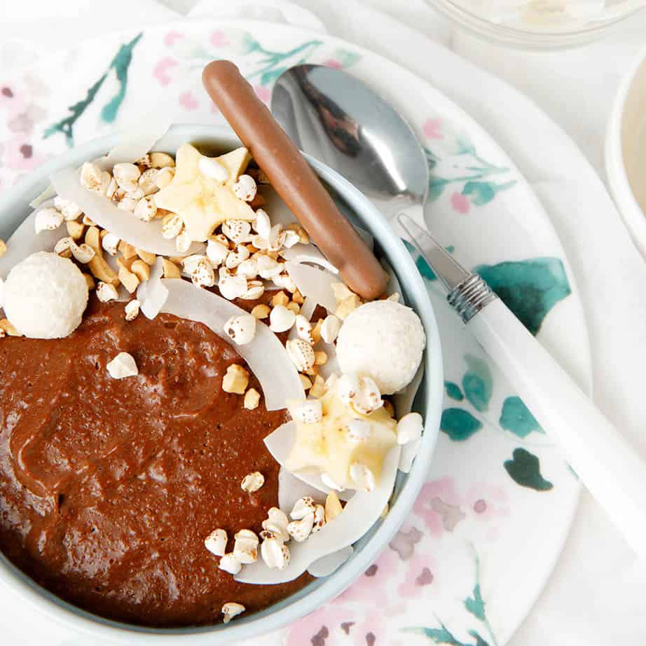 Square image of chocolate oatmeal in a blue bowl topped with bliss balls, banana and nuts