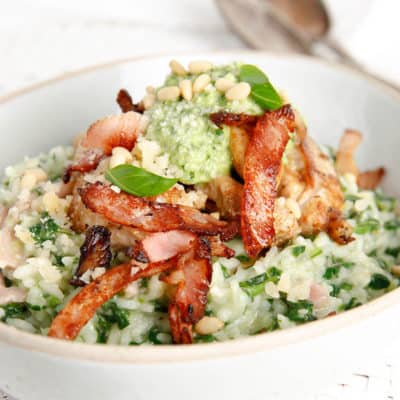 A bowl of chicken risotto topped with pesto and bacon on white background
