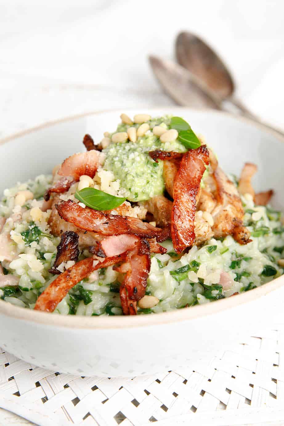 A bowl of chicken risotto topped with pesto and bacon on white background