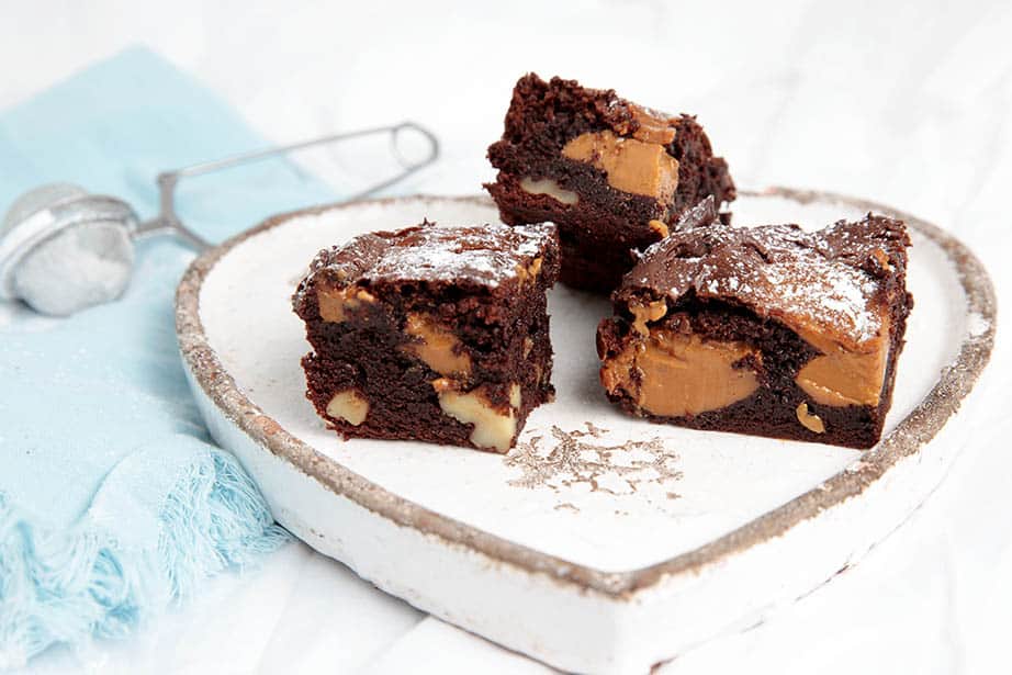 Landscape image of Caramel Chocolate Brownies on a plate and dusted with icing sugar