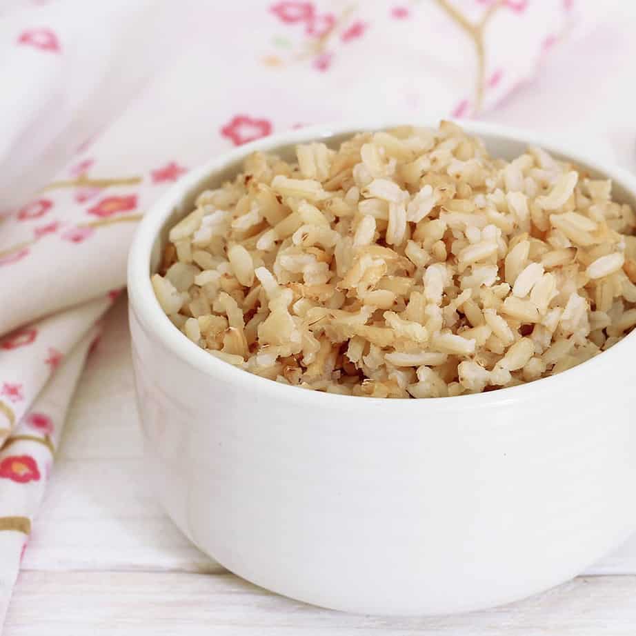 Square crop Cooked brown rice in a white bowl wooden spoon and floral serviette