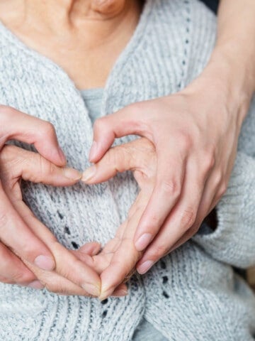 Old and young hands forming a heart on over a ladies heart. SHe is wearing a blue knit jumper.