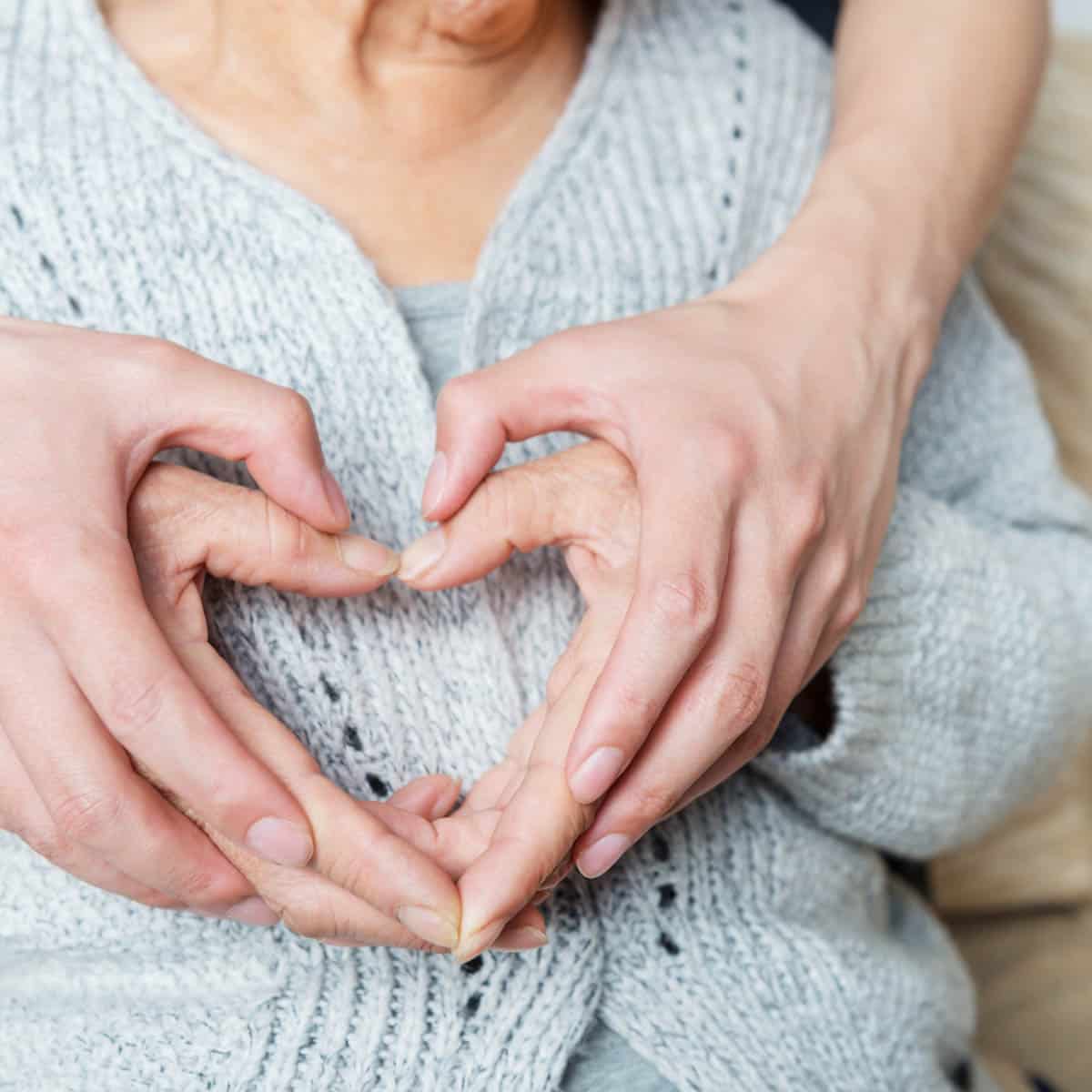 Old and young hands forming a heart on over a ladies heart. SHe is wearing a blue knit jumper.