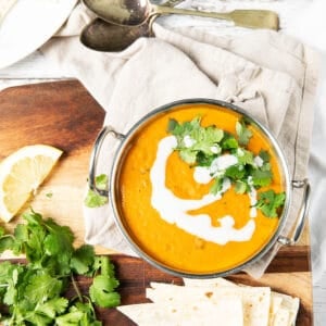 Overhead image of red lentil curry in an Indian serving bowl on a wooden board. Dish served with roti and coriander