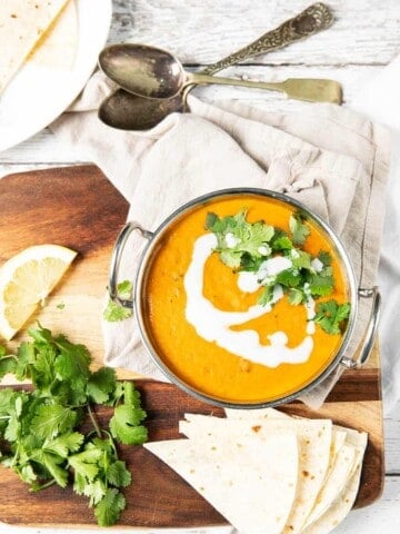 Overhead image of red lentil curry in an Indian serving bowl on a wooden board. Dish served with roti and coriander