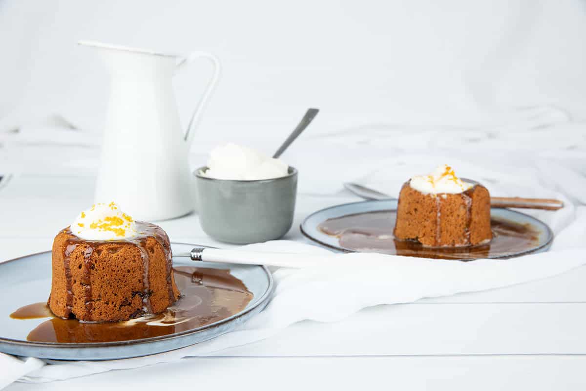 Two Sticky date puddings with caramel sauce in a jug. Cream in a pot and orange zest on white background